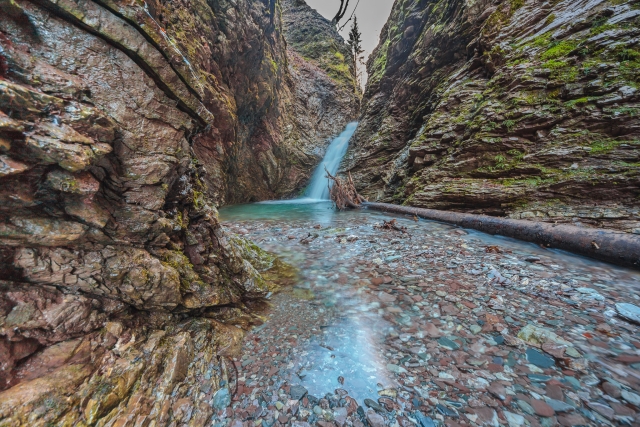 The picture shows a narrow gorge with a river, where a small waterfall cascades. Steep rocky slopes covered with moss and greenery line the river.