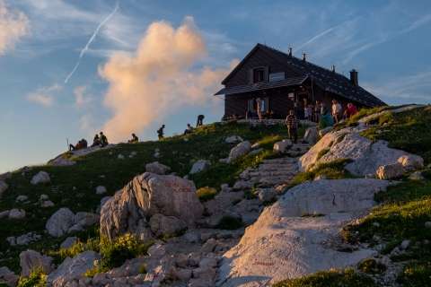 The photograph shows a mountain hut on a mountain peak, with hikers around it. Rocks and grass surround the hut, bathed in the warm light of a sunset.
