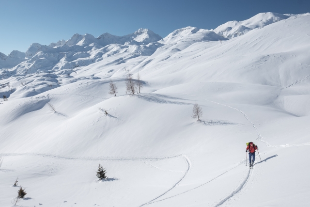Na fotografiji je zasnežena pokrajina s hribi, ki se v ozadju razširijo v višje gorovje. V spodnjem desnem kotu stoji človek, oblečen v polno opremo za turno smučanje, in se vzpenja po snegu.