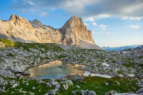 The picture shows a grassy area covered with rocks and a small body of water. In the background, a rocky mountain rises into the sky.