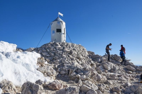 Na fotografiji je planinski vrh z razgledno točko. Nebo je svetlo modro in brez oblakov. V ospredju je gruča sivih in belih kamnov, ki prekrivajo vrh. Na levi strani so ostanki snega, deloma ledeni. V središču je osmerokotna kamnita zgradba s sivo pločevi