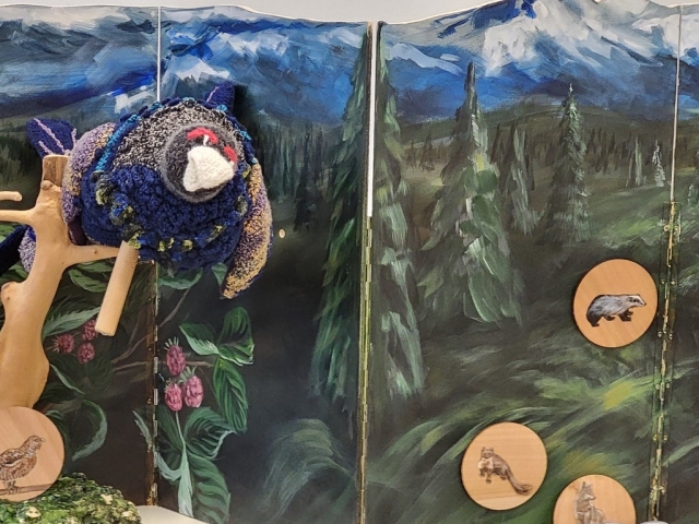 Na fotografiji je iz blaga narejen divji petelin, ki sedi na leseni palici. Za njim je kartonasto ozadje, ki je poslikano z gorami in gozdom. Na njem je tudi pritrjenih nekaj lesenih figuric, na katerih so naslikane različne živali