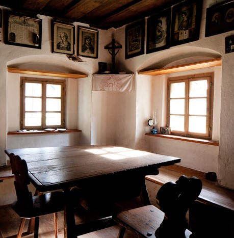 Fotografija prikazuje starinsko jedilnico v svetli sobi s široko leseno mizo in dvema temnima lesenima klopema. Soba ima dva velika okna, ki sta vdelana v obokani zid. Stene so svetle barve, na njih pa visijo slike. Na okenski polici stoji nekaj manjših p