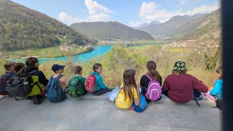 In the photograph there is a group of children sitting on the edge of a slope. Below them are trees, and behind them a landscape with a road, a village, and a blue river on the right, all surrounded by forested hills.