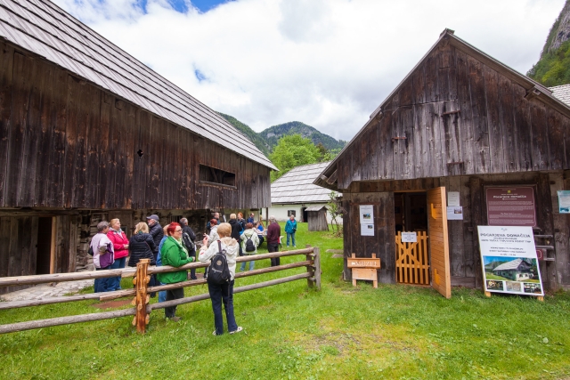 The photo shows two wooden cabins with people standing by them and a wooden fence in front. One cabin has several informational boards. In the background, a stone house and forest-covered hills are visible.
