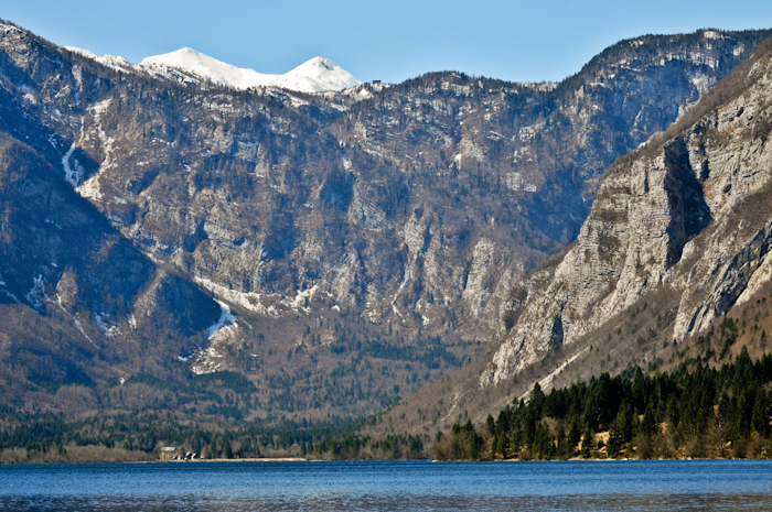 The picture shows the surface of a lake surrounded by wooded and rocky slopes and snow-covered mountains.