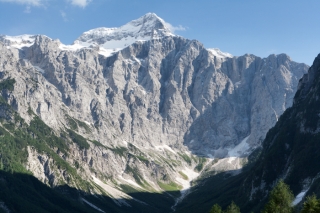 Fotografija prikazuje mogočen gorski vrh, prekrit s snegom, obdan s strmimi skalnatimi pobočji in zelenimi dolinami v spodnjem delu.